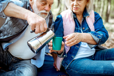 Senior couple pouring coffee having a picnic at the campsite in the forest