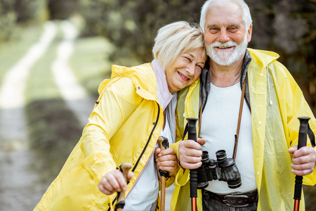 Portrait of a happy senior couple in yellow raincoats hiking with trekking sticks in the young pine forest. Concept of an active lifestyle on retirementの写真素材