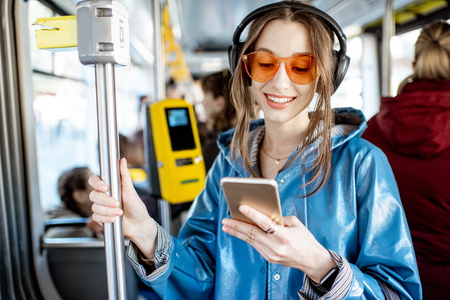 Young stylish woman using public transport, standing with headphones and smartphone while moving in the modern tramの写真素材