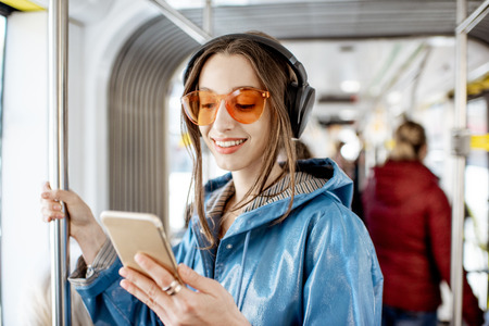 Young stylish woman using public transport, standing with headphones and smartphone while moving in the modern tramの写真素材
