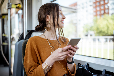 Young and happy woman using smartphone while sitting near the window in the public transport during the tripの写真素材