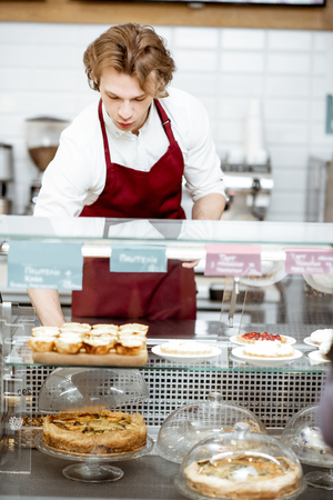 Young salesman in red apron placing sweet pastry in the refrigerator at the showcase of the confectionery shop or cafeの写真素材