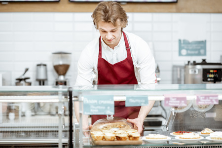 Salesman in red apron putting fresh pie into the refrigerator of the showcase at the modern pastry shopの写真素材