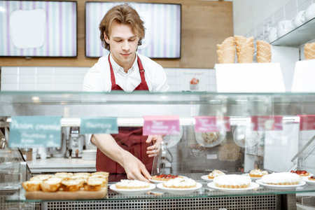 Young salesman in red apron placing sweet pastry in the refrigerator of the showcase at the modern...