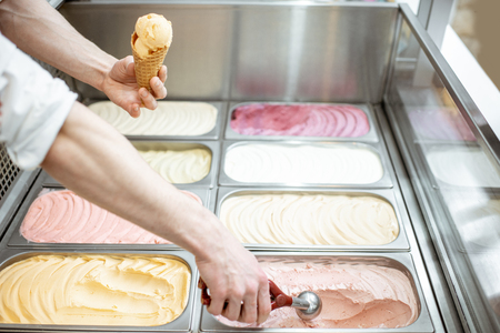 Metal trays full of colorful ice cream in the showcase refrigerator, salesman taking ice cream with scoop, close-up view