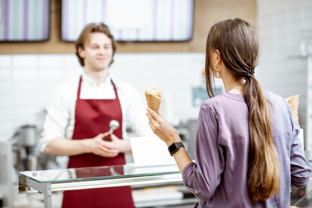 Handsome salesman in red apron making ice cream in waffle cone for a young woman client in the modern pastry shopの写真素材