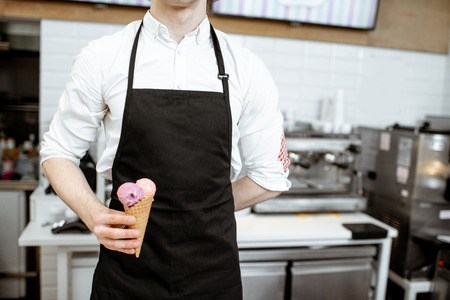Salesman holding ice cream in the waffle cone on the black apron background in the shop, close-up viewの写真素材