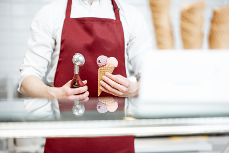 Salesman holding ice cream in the waffle cone and professional scoop on the red apron background, close-up viewの写真素材