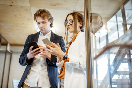 Business man and woman having a conversation while standing together with smart phones in the modern officeの写真素材