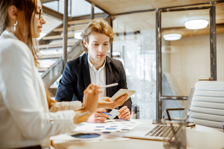 Young man and woman having a serious business conversation, working with paper documents and computer in the officeの写真素材