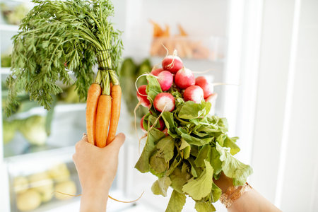 Woman taking fresh radish and carrot from the refrigerator at home, close-up view