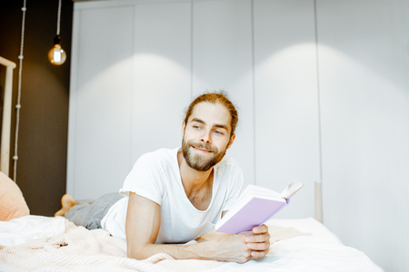 Handsome man in pajamas reading book while relaxing on the bed at homeの写真素材