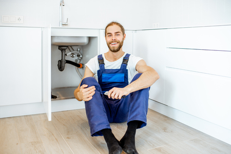 Portrait of a handsome plumber in overalls sitting near the sink at the kitchenの写真素材