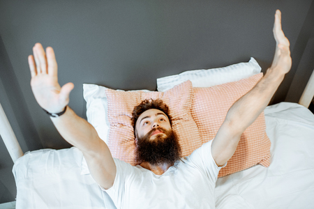 Young bearded man waking up, lying on the bed with raised handsの写真素材
