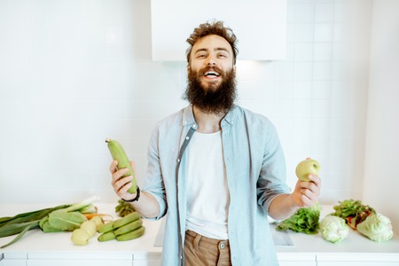 Portrait of a handsome bearded man standing with fresh green vegetables and fruits on the kitchenの写真素材