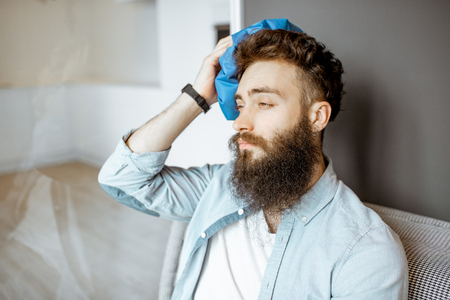 Bearded man with headache applying ice bag while sitting on the couch at homeの写真素材