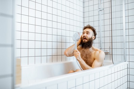 Joyful bearded man washing in the bathtub, having fun singing into the shower in the bathroomの写真素材