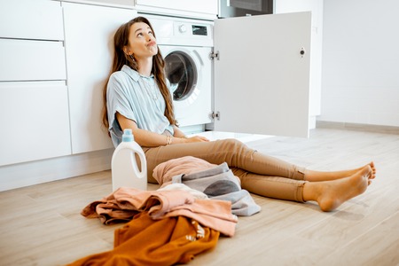 Portrait of a young and thoughtful woman sitting on the floor, washing clothes at homeの写真素材