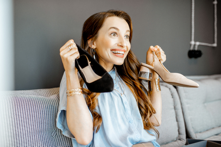 Portrait of a young woman excited with a new purchases, holding beautiful shoes while sitting on the couch at homeの写真素材