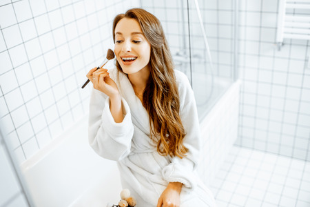 Beautiful woman taking care of herself, applying some cosmetics while sitting on the bathtub in the white bathroom interiorの写真素材