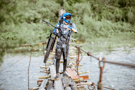 Professional well-equipped cyclist carrying bicycle on the old wooden bridge in the mountains....