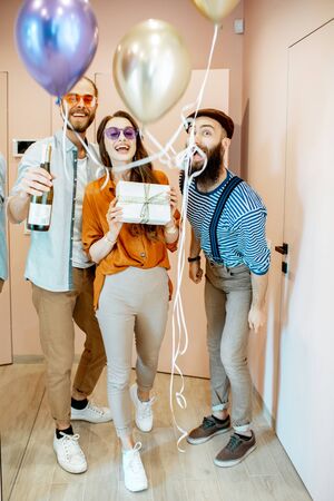 Joyful friends coming with balloons, gifts and drink for the celebration, standing in the apartment hall near the entranceの写真素材