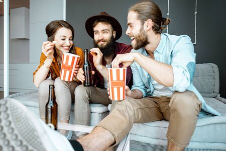 Young friends watching some movie, sitting together with pop cornes on the couch at homeの写真素材