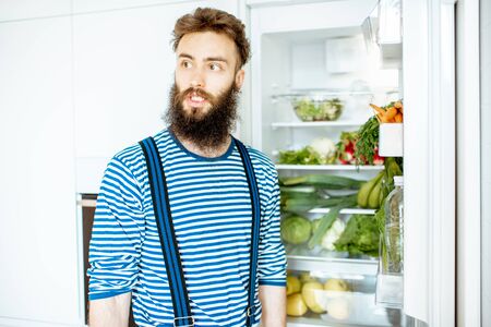 Portrait of a well-looking man standing near the fridge full of fresh vegetables and fruits at homeの写真素材