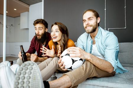 Young friends watching football match on the tv, cheering up for a favorite team while sitting together on the couch at homeの写真素材