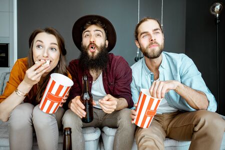 Young friends watching some movie, sitting together with pop cornes on the couch at homeの写真素材