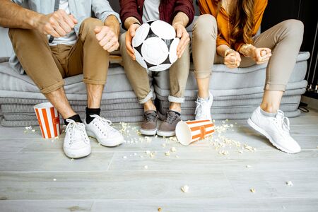 Friends watching football match. View on their legs with pop cornes and ball on the floorの写真素材