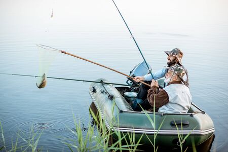 Grandfather with adult son fishing on the inflatable boat, catching fish with net on the lake early in the morningの写真素材