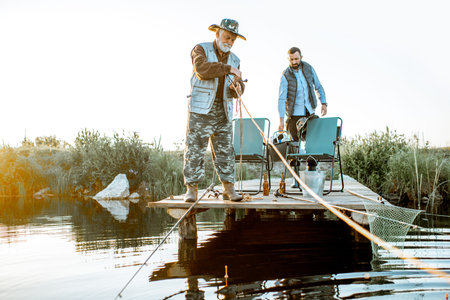 Grandfather with adult son preparing for fishing, standing on the wooden pier during the morning...