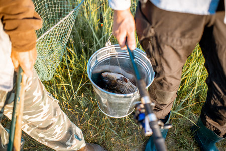 Men holding fishing rods and bucket with fresh caught fish outdoors. Close-up view with no face
