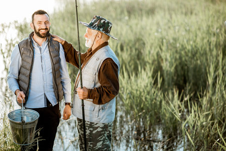 Portrait of a senior grandfather with adult son fishing on the lake, standing together between the reeds during the morning lightの写真素材
