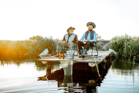 Grandfather with adult son fishing together on the wooden pier during the morning light. View from...