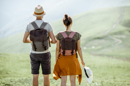Young couple standing together with backpacks, enjoying beautiful view on the green mountains, rear...