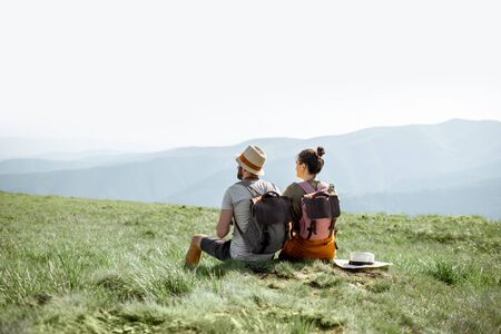 Young couple sitting on the green meadow, traveling with backpacks in the mountains during the summer timeの写真素材