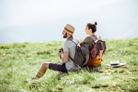 Young couple sitting on the green meadow, traveling with backpacks in the mountains during the summer timeの写真素材