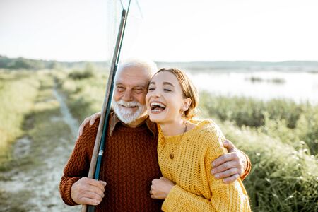 Portrait of a young woman with cheerful senior grandfather hugging together outdoors, having a good time while fishingの写真素材