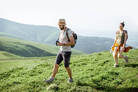 Beautiful couple walking with backpacks on the green meadow, having fun while traveling in the mountains during the summer timeの写真素材