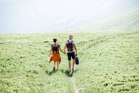 Couple walking with backpacks on the green meadow, traveling in the mountains during the summer time, wide landscape viewの写真素材