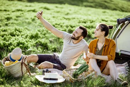 Young and cheerful couple making selfie photo during the picnic at the campsite, traveling high in the mountains during the sunsetの写真素材