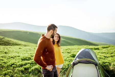 Lovely couple in bright sweaters walking on the green meadow, while traveling with tent high in the mountains during the sunsetの写真素材