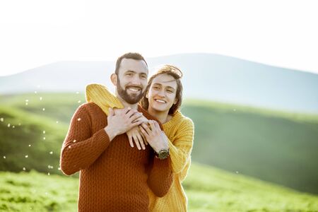 Portrait of a lovely couple dressed in bright sweaters hugging on the green meadow while traveling in the mountainsの写真素材