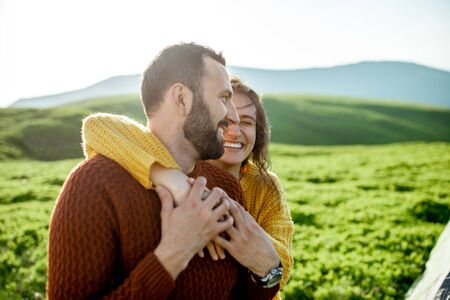 Lovely couple in bright sweaters hugging together on the green meadow, while traveling high in the mountains during the sunsetの写真素材