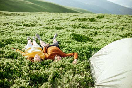Lovely couple in bright sweaters lying on the green meadow near the campsite in the mountainsの写真素材