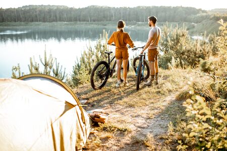 Young couple standing with mountain bicycles at the campsite, traveling in the forest near the lake on the sunsetの写真素材