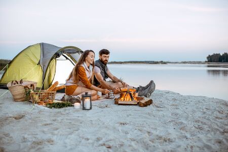 Young and cheerful couple sitting at the fireplace, cooking sausages, having a picnic at the campsite on the beach in the eveningの写真素材
