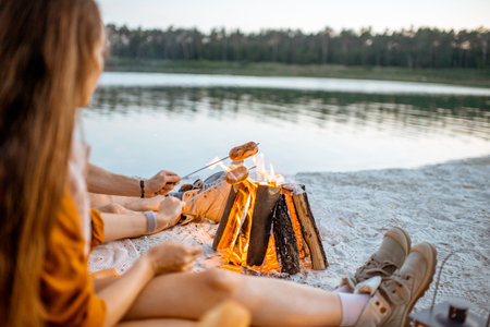 Couple cooking sausages at the fireplace, having a picnic at the campsite on the beach in the...